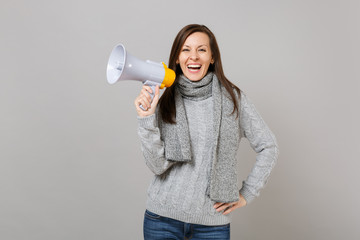 Laughing young woman in gray sweater, scarf holding megaphone isolated on grey background, studio portrait. Healthy fashion lifestyle, people sincere emotions, cold season concept. Mock up copy space.