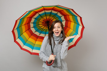 Surprised young woman in gray sweater, scarf spreading hands, holding colorful umbrella isolated on...