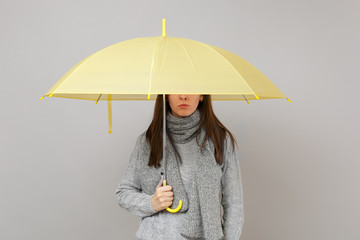 Young woman in gray sweater, scarf holding yellow umbrella, covering eyes isolated on grey wall background. Healthy fashion lifestyle, people sincere emotions, cold season concept. Mock up copy space.