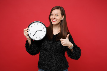 Portrait of stunning young woman in black fur sweater showing thumb up, holding round clock...