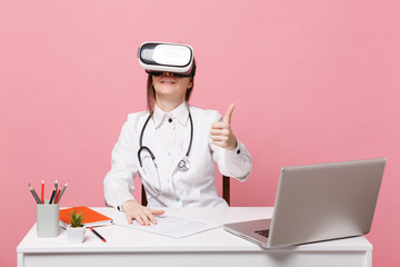 Female doctor sit at desk work on computer with medical document in headset in hospital isolated on pastel pink wall background. Woman in medical gown glasses stethoscope. Healthcare medicine concept.