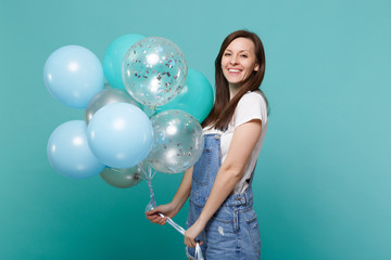 Fototapeta premium Portrait of pretty smiling young happy woman in denim clothes celebrating and holding colorful air balloons isolated on blue turquoise wall background. Birthday holiday party, people emotions concept.