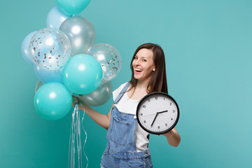 Portrait of cheerful young woman in denim clothes holding round clock and celebrating with colorful...