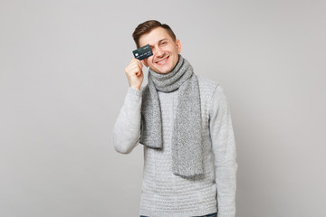 Cheerful young man in gray sweater, scarf covering eye with credit bank card isolated on grey background. Healthy fashion lifestyle, people sincere emotions, cold season concept. Mock up copy space.