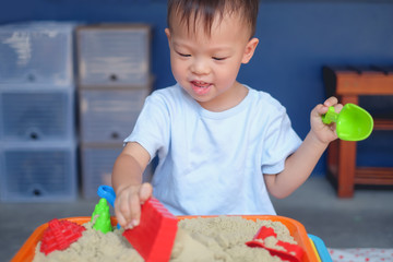 Cute smiling Asian 2 - 3 years old toddler boy playing with kinetic sand in sandbox at home /...