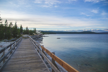 Fototapeta na wymiar Wooden pier on the beach of Yellowstone lake. Mountains view on background. Small forest on the side. Blue sky with some clouds and blue lake with geysers. 