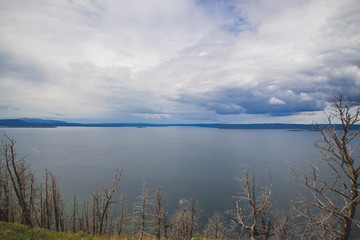 The Yellostone lake  with clouds. Blue sky and rainy clouds on the top. Hills view. Trees with green grass in front of lake. 