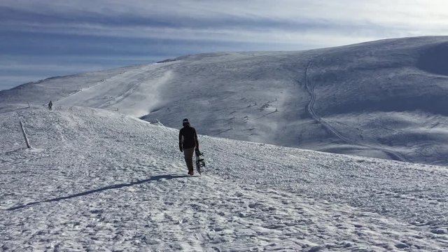  Snowboarder walks on top of a mountain against the backdrop of a mountain view while holding a board in his hand.