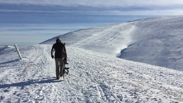  Snowboarder walks on top of a mountain against the backdrop of a mountain view while holding a board in his hand.