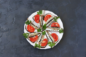Sandwiches with sprats and tomatoes on a white plate on a dark background, top view