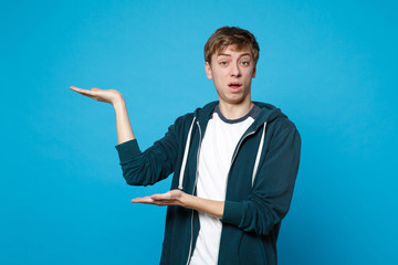 Portrait of perplexed young man in casual clothes pointing hands aside isolated on blue wall background in studio. People sincere emotions, lifestyle concept. Mock up copy space. Advertising area.