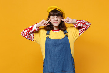 Girl teenager in french beret denim sundress keeping eyes closed covering ears with fingers isolated on yellow wall background in studio. People sincere emotions lifestyle concept. Mock up copy space.