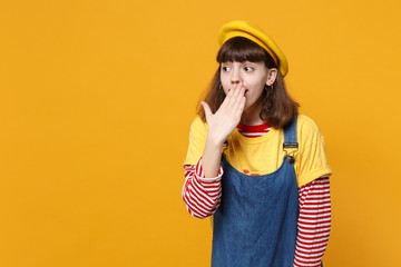 Amazed girl teenager in french beret, denim sundress covering mouth with hand looking aside isolated on yellow wall background in studio. People sincere emotions lifestyle concept. Mock up copy space.