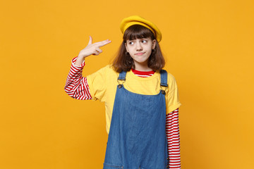 Upset girl teenager in french beret, denim sundress point fingers to head as if she about shoot herself isolated on yellow background. People sincere emotions, lifestyle concept. Mock up copy space.