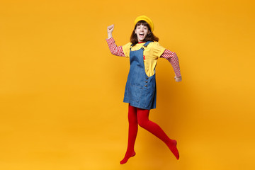 Cheerful girl teenager in french beret, denim sundress spreading hands, jumping and fooling around isolated on yellow wall background in studio. People emotions, lifestyle concept. Mock up copy space.