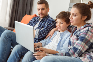 Fototapeta premium Family at home sitting on couch in living room together browsing internet on laptop concentrated