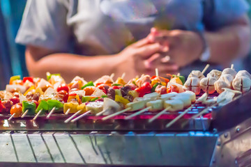 Seafood and beef barbecue (BBQ) skewered on bamboo sticks with pie apple, tomato, onion and chili are grilling on the stove for barbecue party. Barbecue skewers meat kebabs with vegetables at night.