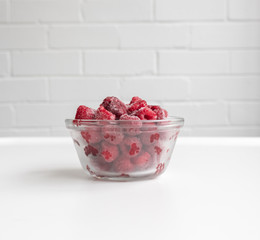 Close up of frozen red raspberries in small glass dish on white table (selective focus)