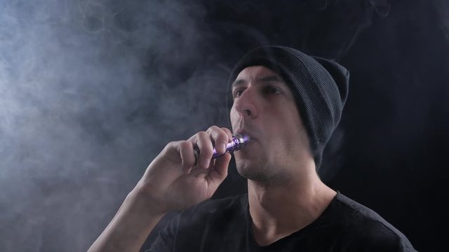 young man vaper exhaling big clouds of smoke with e-cigarette vape on black background in slow motion