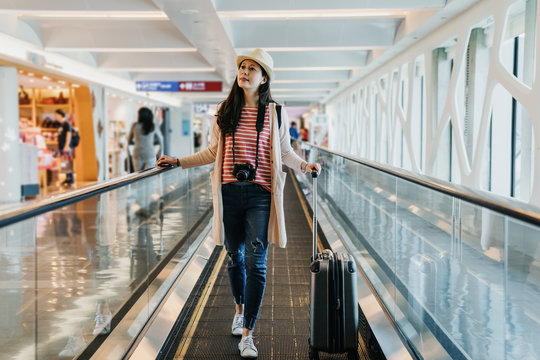 asian tourist elegant woman walking on the moving walkway at the airport with black suitcase. young girl traveler in hat carrying luggage pass tax free zone window shopping in duty free area.