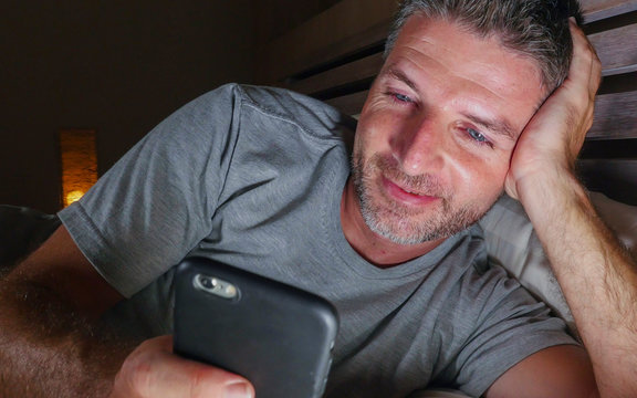 young handsome and attractive happy man networking with mobile phone late at night  in bed smiling relaxed enjoying social media or online dating app in internet addiction
