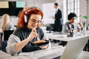 Smiling businesswoman eating salad while listening music on headphones at work.