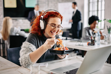 Cheerful businesswoman eating salad and listening music on headphones in the office.