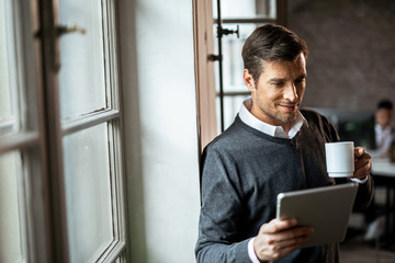 Smiling businessman drinking coffee and surfing the net on touchpad by the window.