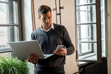 Businessman reading text message on mobile phone while using laptop by the window.