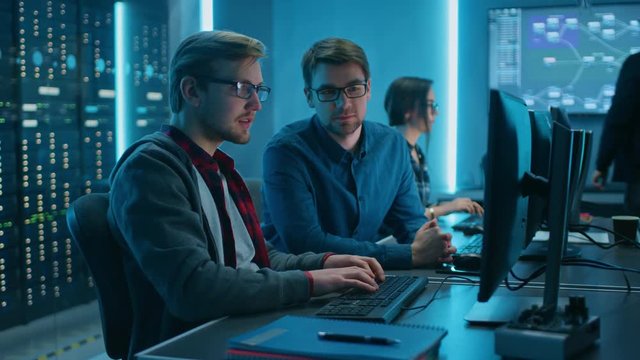 Two Professional IT Programers Discussing Technical Data Show on Desktop Computer Display. Working Data Center Technical Department with Server Racks in the Background