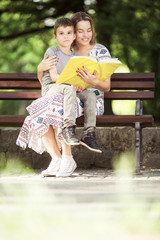 Mother and son sitting on a bench outdoors in park and reading a book