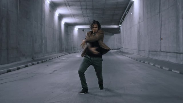 Serious Cool Young Hipster Man with Long Hair is Energetically Dancing Hip Hop in a Lit Concrete Tunnel. He's Wearing a Brown Leather Jacket.