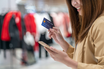 Closeup Asian woman using credit card with mobile for online shopping in department store over the clothes shop store background, technology money wallet and online payment concept, credit card mockup