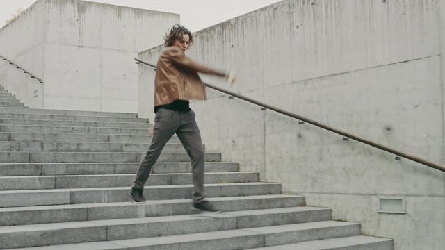 Cheerful and Happy Young Man with Long Hair Actively Dancing While Walking Down the Stairs. He's Wearing a Brown Leather Jacket. Scene Shot in an Urban Concrete Park. Day is Bright.