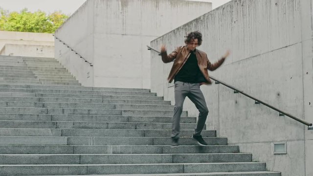 Cheerful and Happy Young Man with Long Hair Actively Dancing While Walking Down the Stairs. He's Wearing a Brown Leather Jacket. Scene Shot in an Urban Concrete Park. Day is Bright.