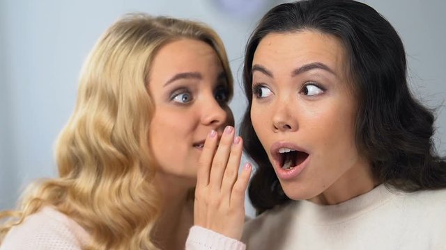 Pretty blonde girl whispering secrets to her attractive asian friend, gossips
