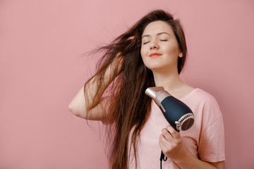 young woman makes hair volume with  hairdryer in hand on pink background