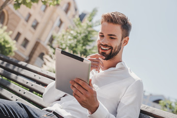 Social life. Young smiling man sitting on a bench in the park, holding his tablet. Rest, communication and relax concept