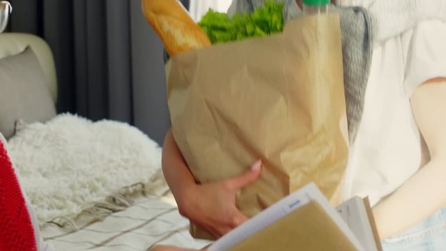 Young Woman Bringing Paper Bag With Groceries And Giving It To Senior Grandmother While Helping Her At Home