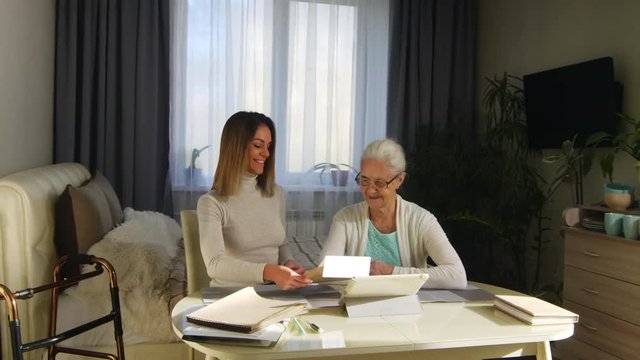 Young cheerful woman bringing a letter to senior grandmother while watching family pictures and talking at table in home