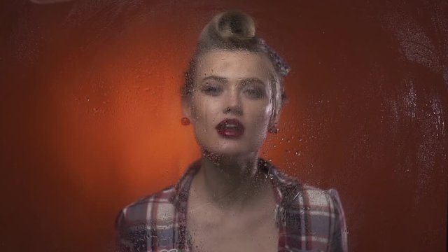 Sexy retro blonde is behind the wet window and spraying it with water