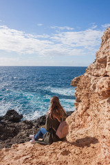 Happy female tourist in coat sits on cliff and enjoys sea view in Malta