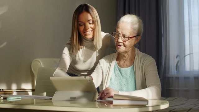 Young Caucasian woman smiling and explaining senior grandmother how to use new digital tablet while spending a time together at home