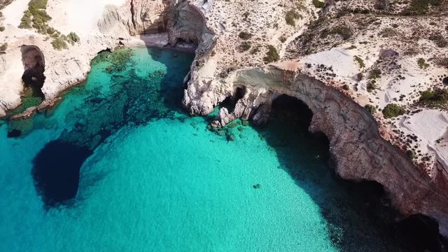 Aerial drone bird's eye view video of iconic volcanic white chalk beach and caves of Tsigrado with turquoise and sapphire clear waters, Milos island, Cyclades, Greece