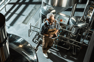 overhead view of brewer in working overalls using digital tablet in brewery - Powered by Adobe