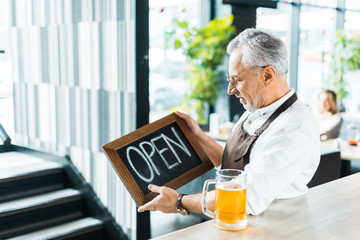 senior male owner of pub holding open sign and standing near bar counter with glass of beer