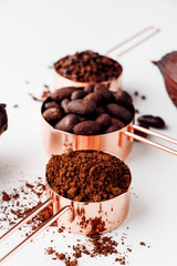 Rose gold measuring cups of cocoa beans, cacao nips, cocoa powder and cocoa pods on a white background, healthy food concept