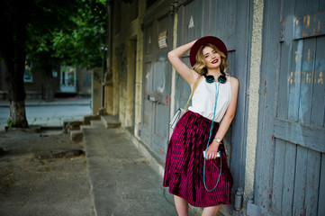 Fashionable and beautiful blonde model girl in stylish red velvet velour skirt, white blouse and hat, posed with phone and earphones.