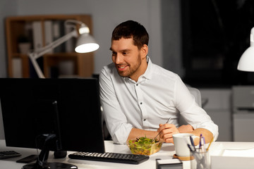 business, overwork, deadline and people concept - smiling businessman at computer eating vegetable salad at night office