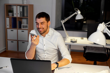 business, deadline and technology concept - smiling businessman with laptop computer using voice command recorder on smartphone at night office
